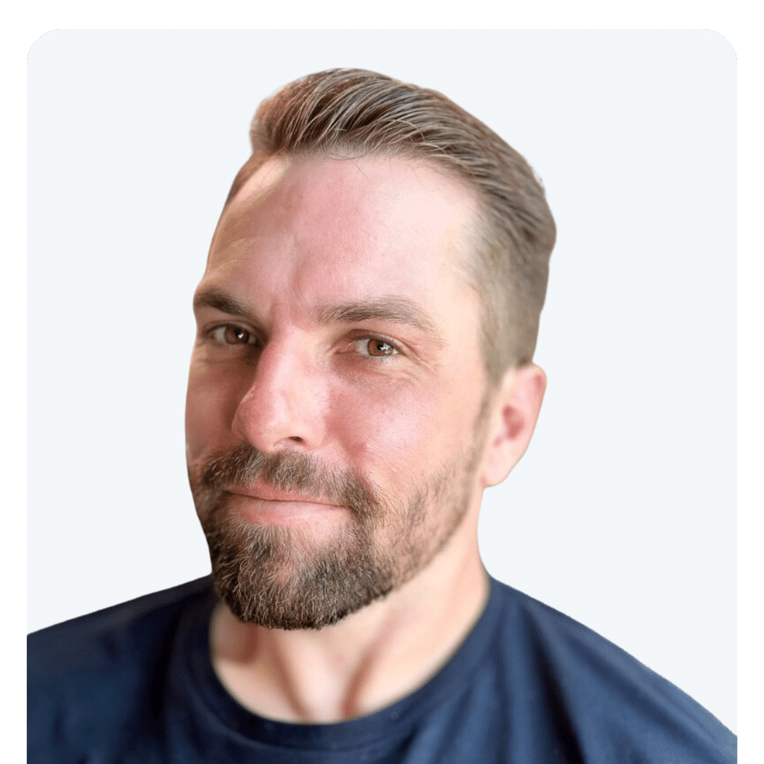 A man with short hair and a beard is looking into the camera, smiling slightly. He is wearing a dark t-shirt and has a light-colored background, resembling someone engaged in market research or data collection.