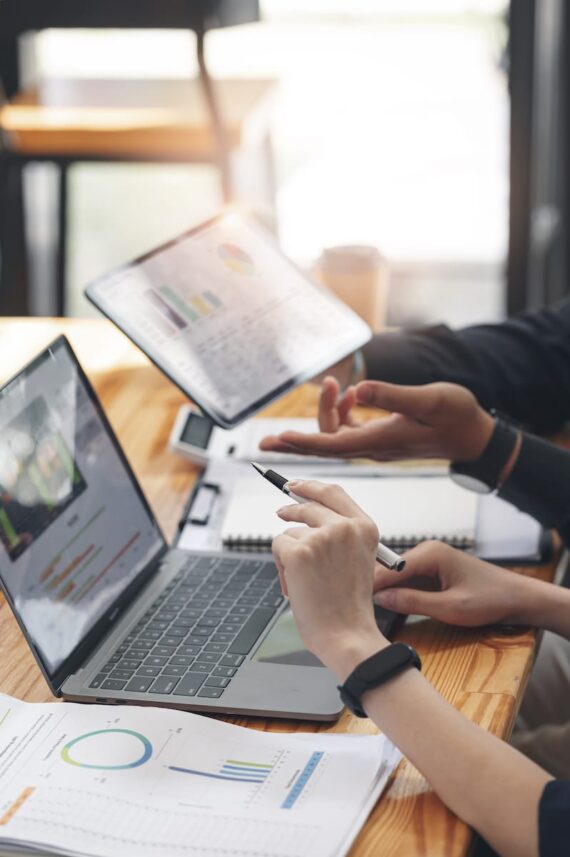 Two people sit at a wooden table with laptops, tablets, and notebooks. Both focus on market research, working with charts and graphs; one gestures while the other, a survey programmer, points at a tablet with a stylus for data collection.