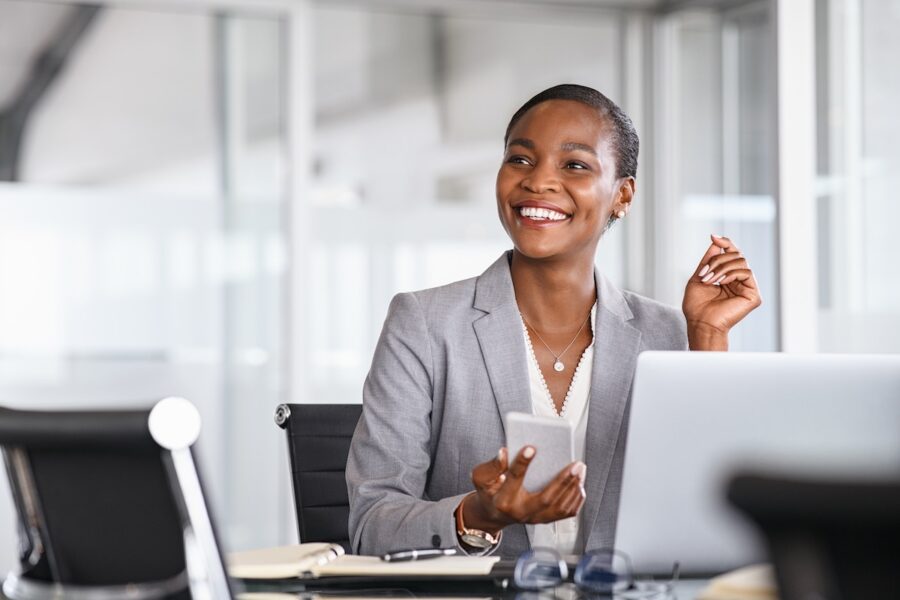 A person in a gray suit is sitting at a desk with a laptop, holding a smartphone and smiling, likely reviewing market research. The background is an office setting with large windows.