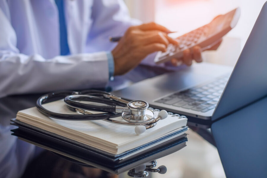 stethoscope on a notebook next to a laptop while someone in a lab coat types on a calculator