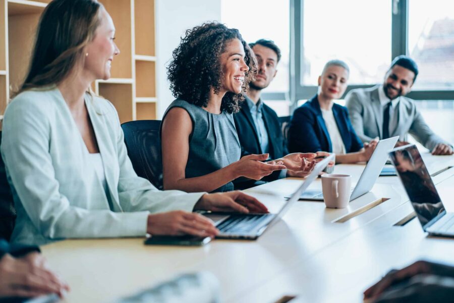 woman smiling gesturing and talking during a corporate meeting surrounded by colleagues on laptops.